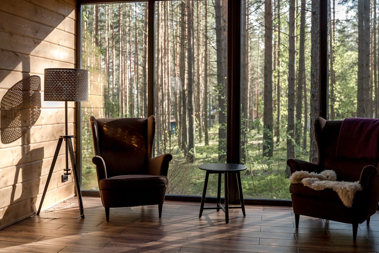 Serene living room with armchairs and lamp, overlooking a lush forest through large windows.